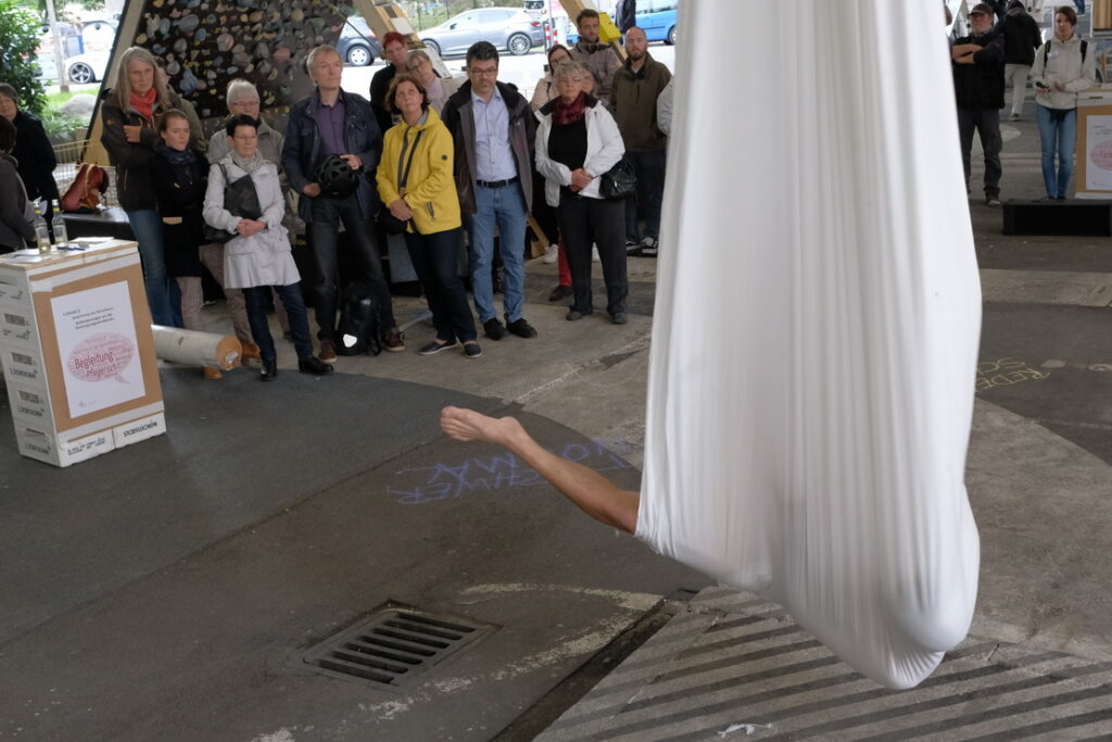 Bild: Künstler in Segeltuch hängt von der Brücke und streckt ein Bein raus